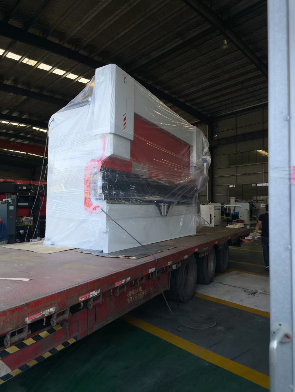 Front three-quarter view of a 130T/4000 press brake wrapped in stretch film on a flatbed trailer inside the ZTGAOKE workshop, with the Y1 cylinder housing visible above the red side panel