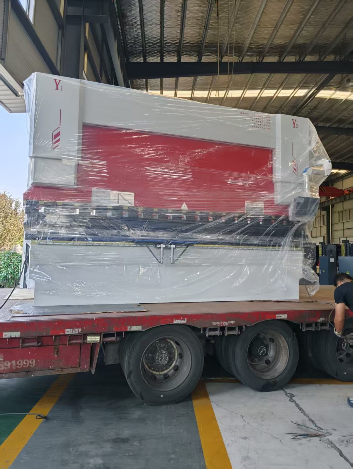 Side view of the stretch-wrapped 130T/4000 press brake on the trailer showing the full 4000 mm bed, the Y1 and Y2 cylinder housings at both ends and the CNC control pendant wrapped at the right side